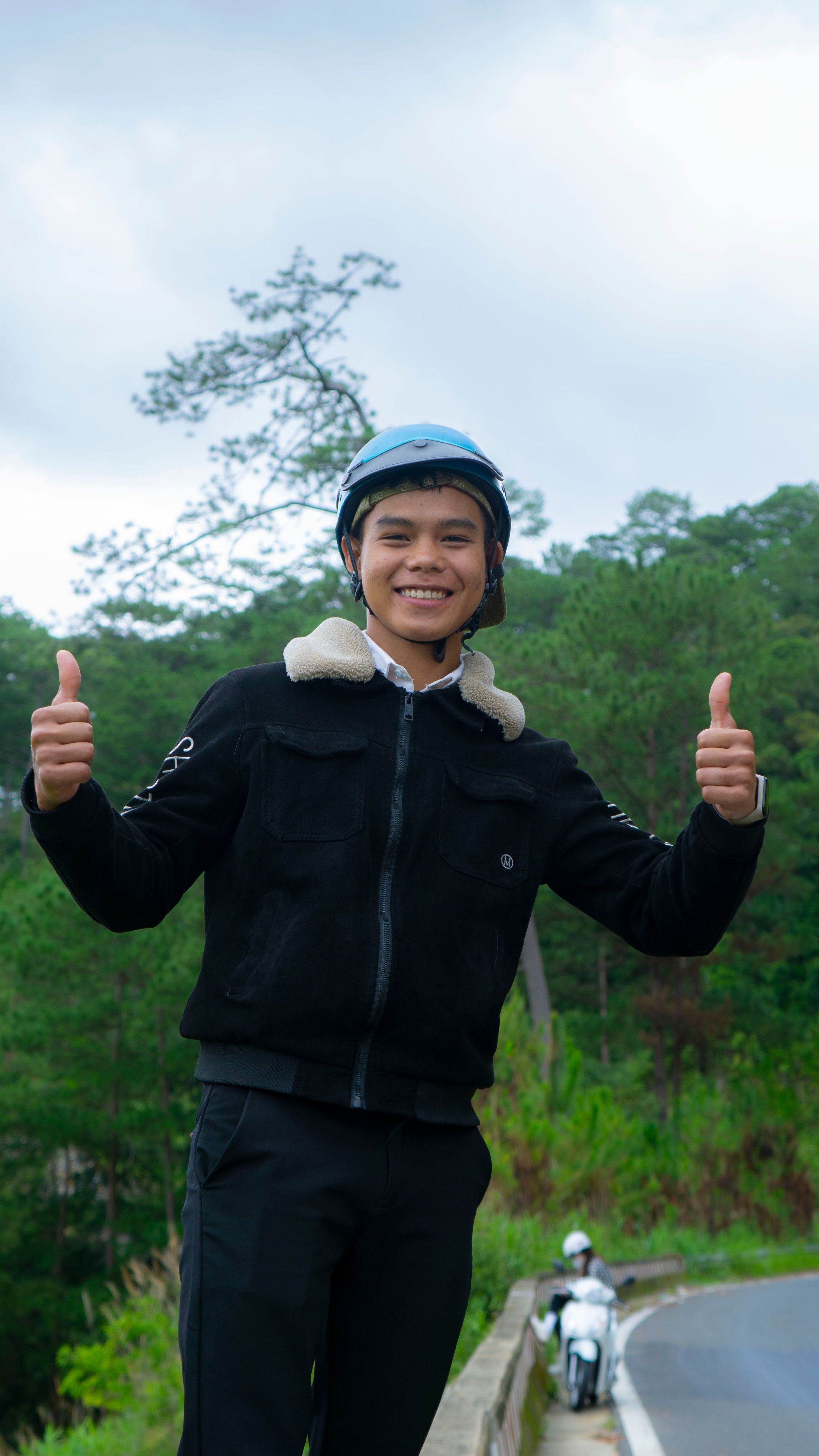 Smiling Asian teenager in helmet giving thumbs up outdoors on a scenic road.