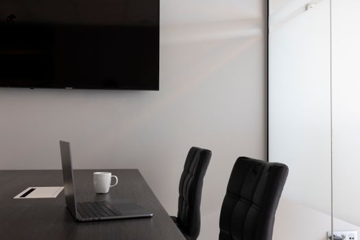 A sleek, modern office setup with a laptop, cup, and TV in an empty conference room.