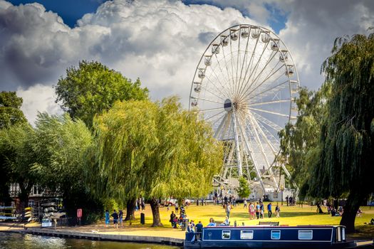 Captured in Stratford-upon-Avon, the Ferris wheel stands tall against a cloudy sky, surrounded by vibrant greenery and lively park-goers.