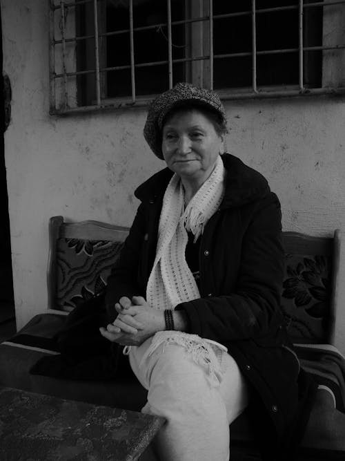Grayscale Photo of a Woman in Black Coat with White Scarf Sitting on Bench