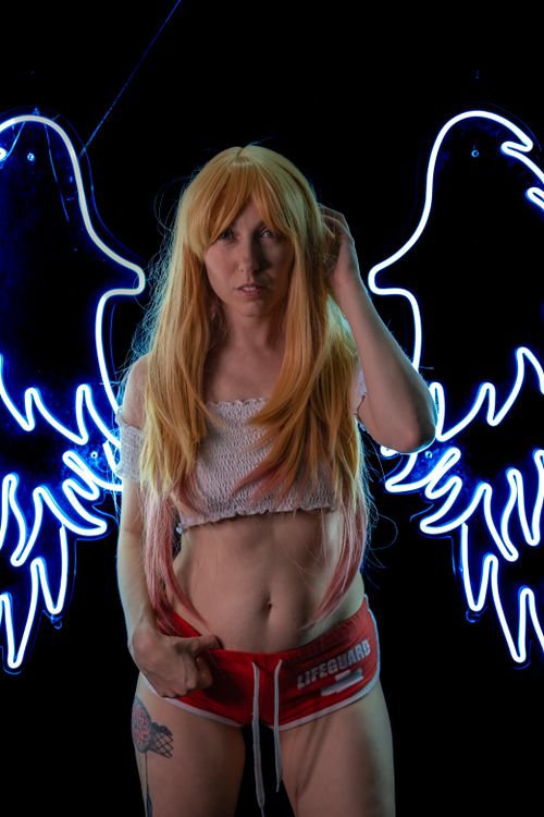 Free Creative portrait of a blonde woman posing in front of neon angel wings at night. Stock Photo
