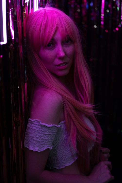 Free A woman with vibrant pink hair poses in neon lighting, wearing a white off-shoulder top. Stock Photo