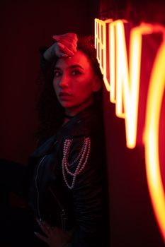 A fashionable woman poses in dim lighting with vibrant neon signs, adding a modern and artistic vibe.