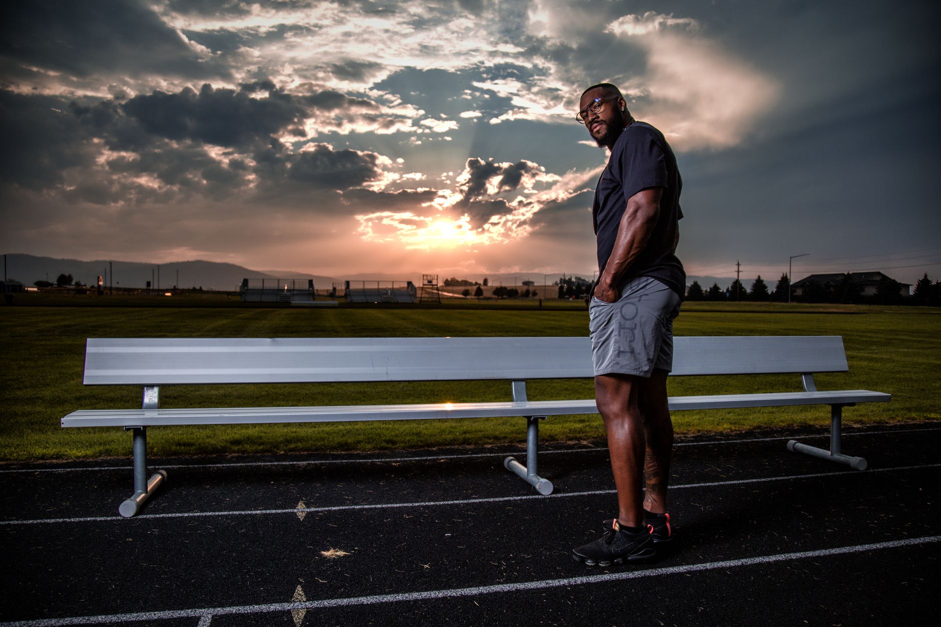 A man in activewear stands beside a bench on a track, with a scenic sunset sky in the background.