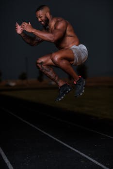 A fit, shirtless man executing a power jump on a dark outdoor track, showcasing athleticism.