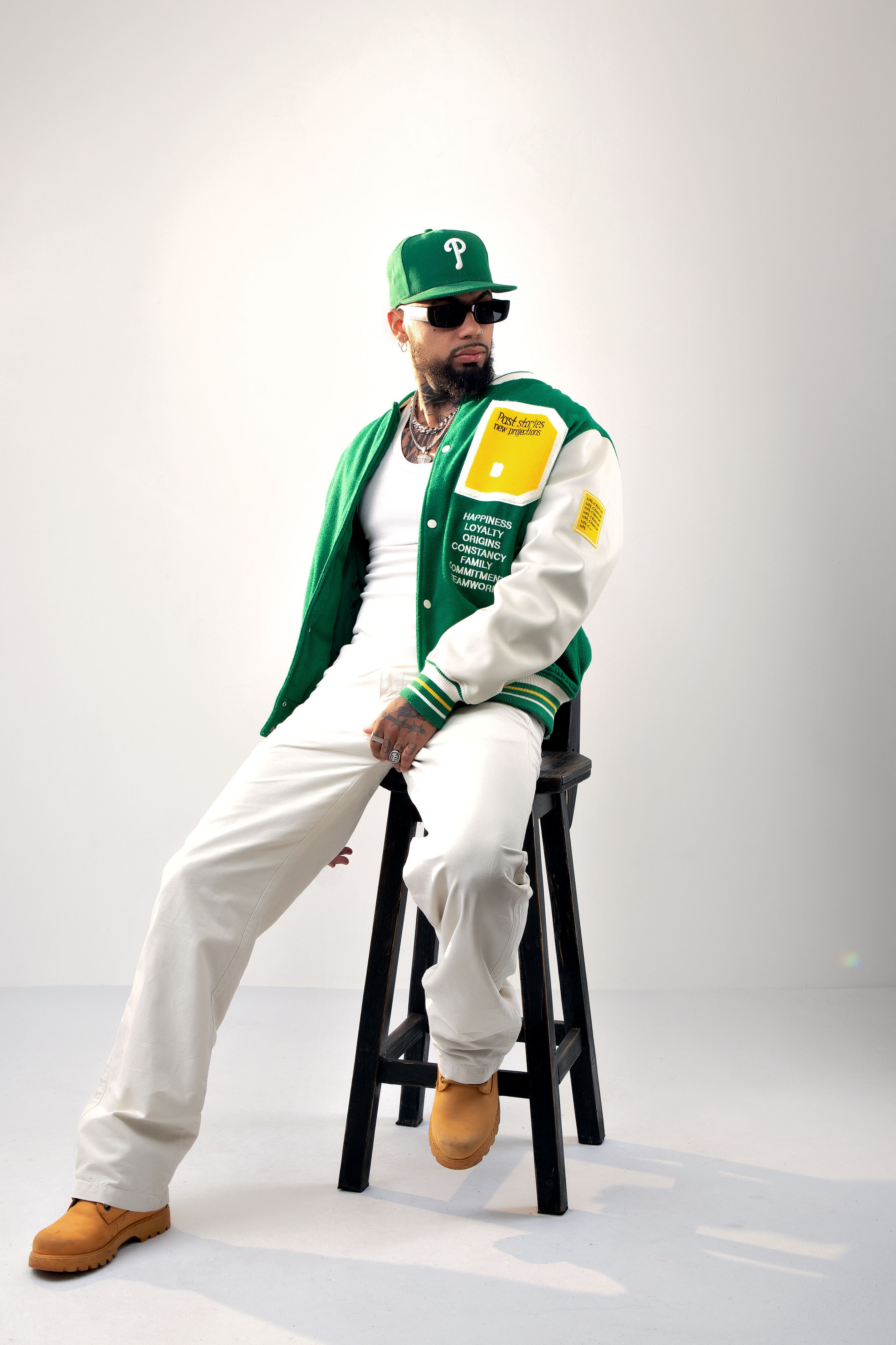 Fashionable man in sunglasses and cap sitting on stool in studio setting.