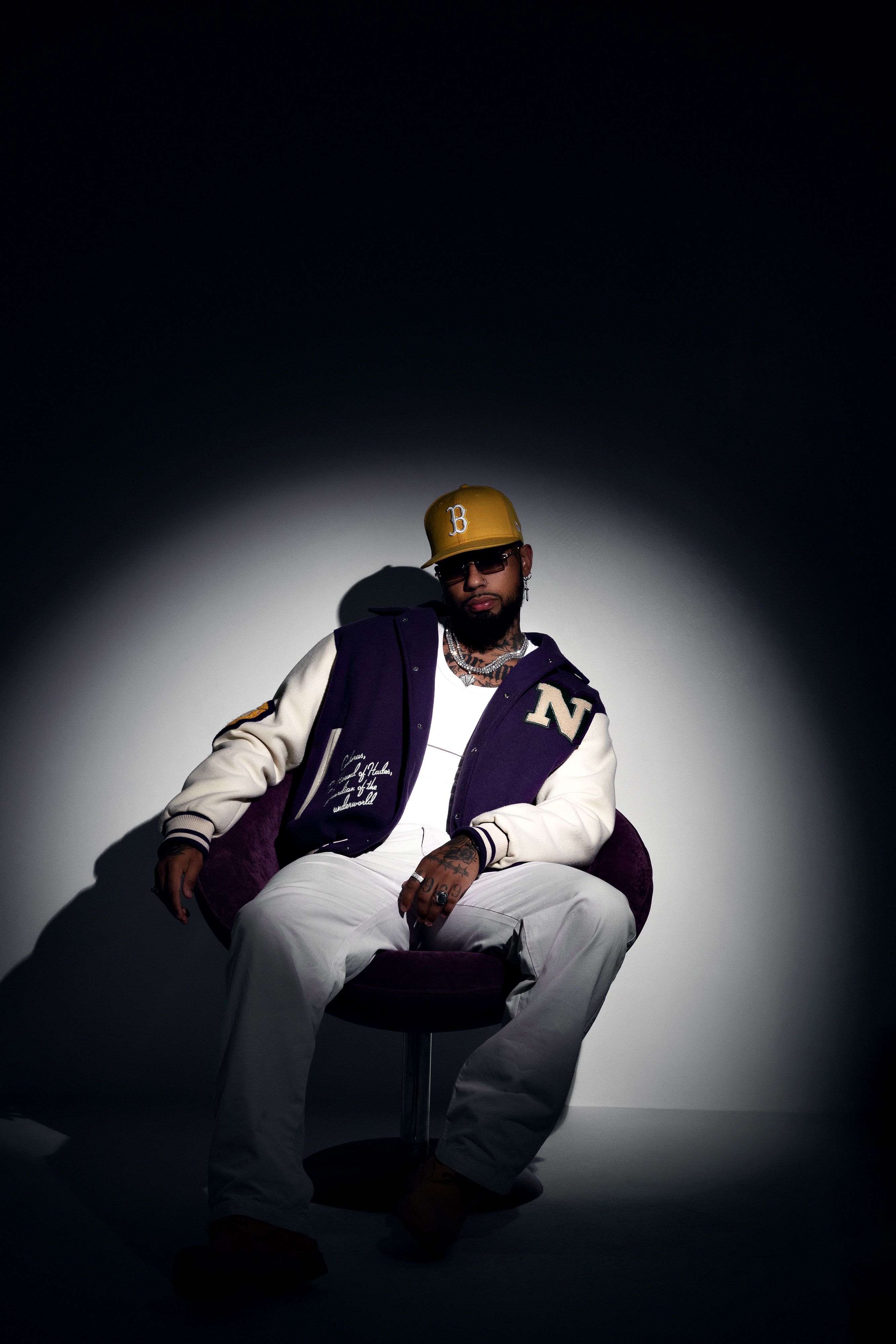 Man with sunglasses and jacket sitting on a chair in a dark studio setting.