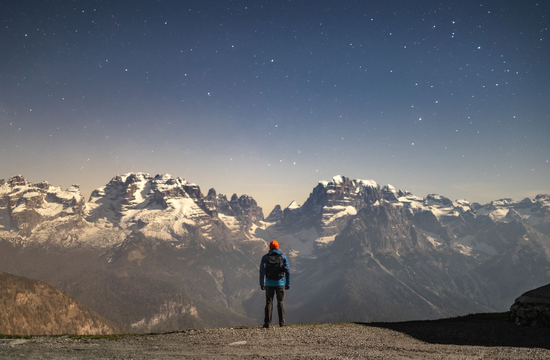 A lone hiker admires a breathtaking snow-capped mountain range under a starry night sky.