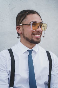 Bearded man with sunglasses, tie, and suspenders, smiling stylishly against a neutral background.