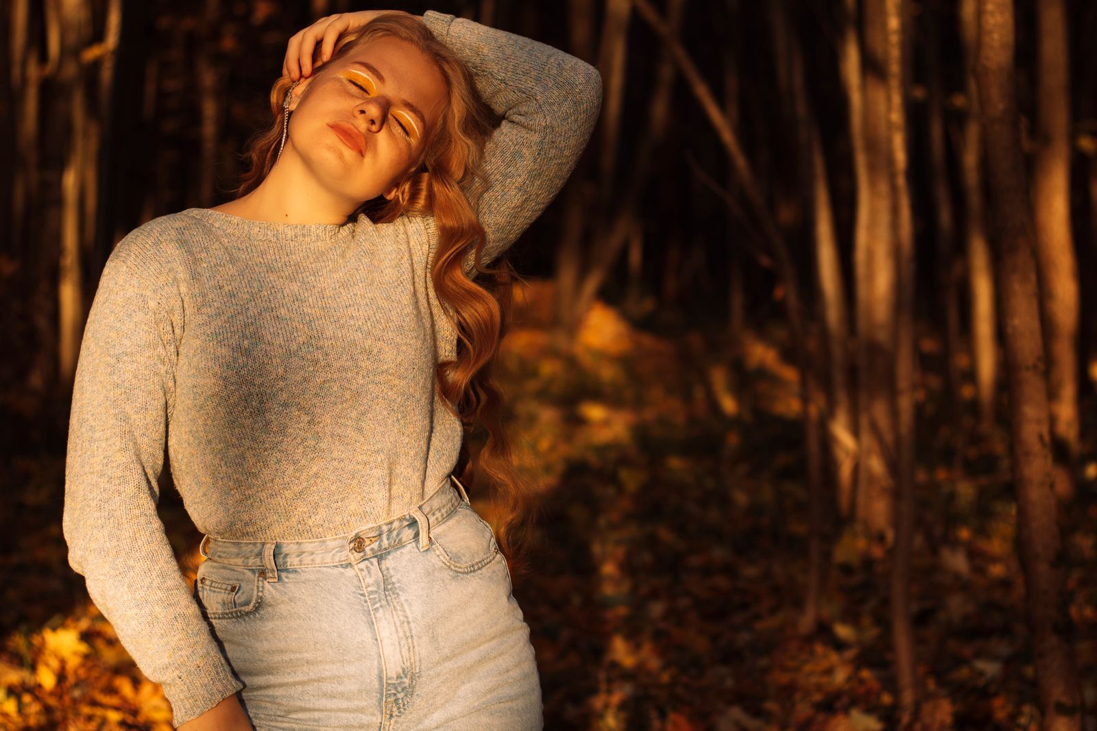 Woman with long wavy hair in a gray sweater, eyes closed, standing in golden autumn forest light