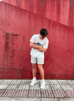 Young man stands with arms crossed, looking down in casual attire against a bold red wall.
