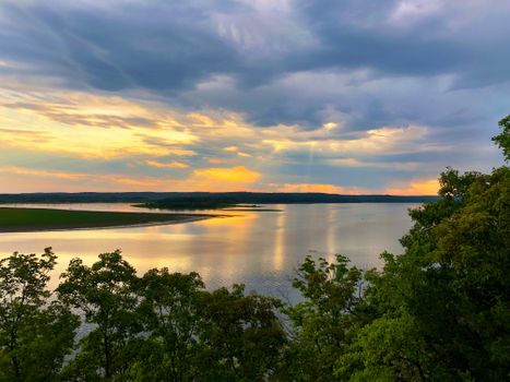 A peaceful sunset reflecting on a tranquil river surrounded by trees and cloudy sky.