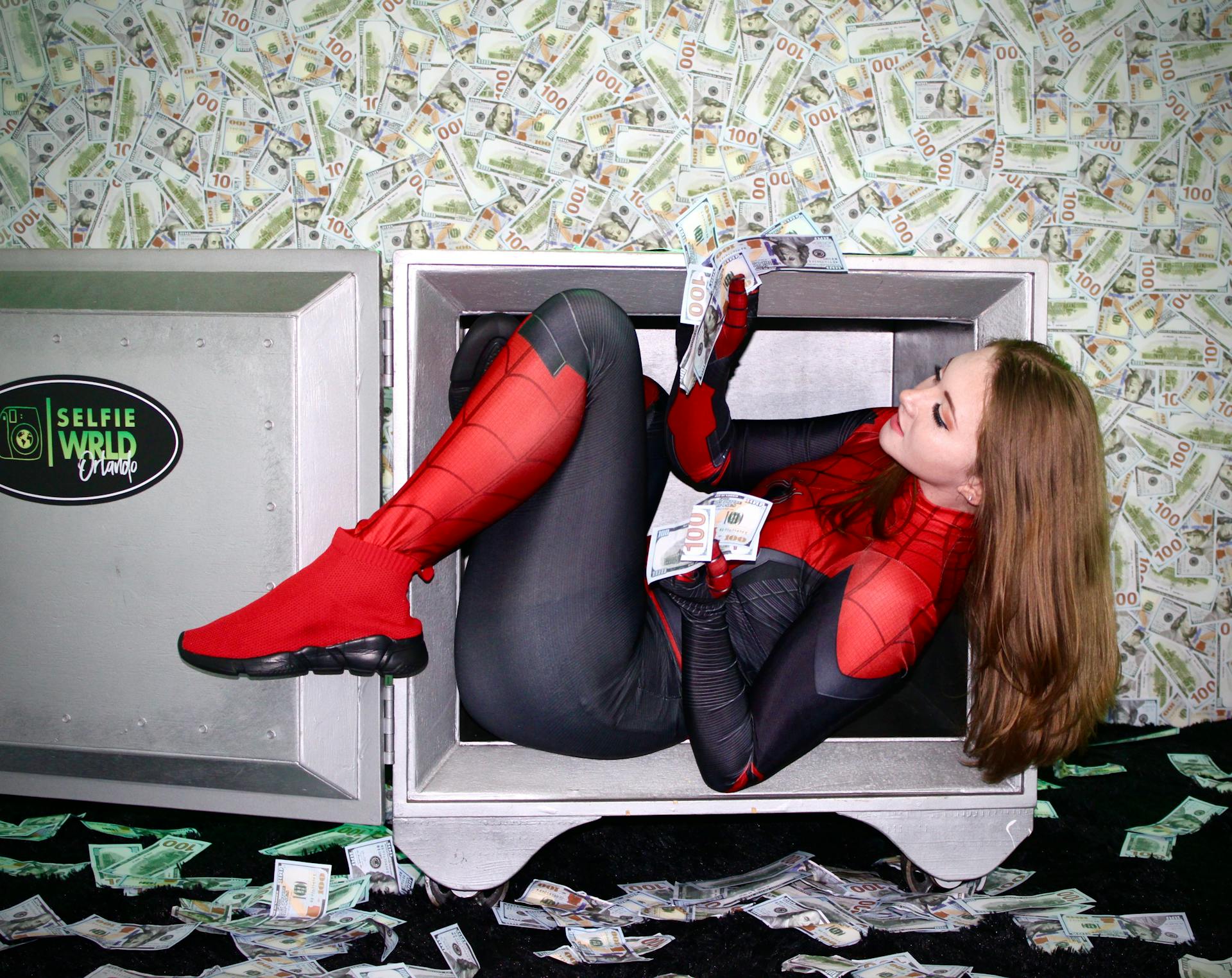 Young woman in Spiderman costume inside a vault, surrounded by cash for a playful photoshoot.