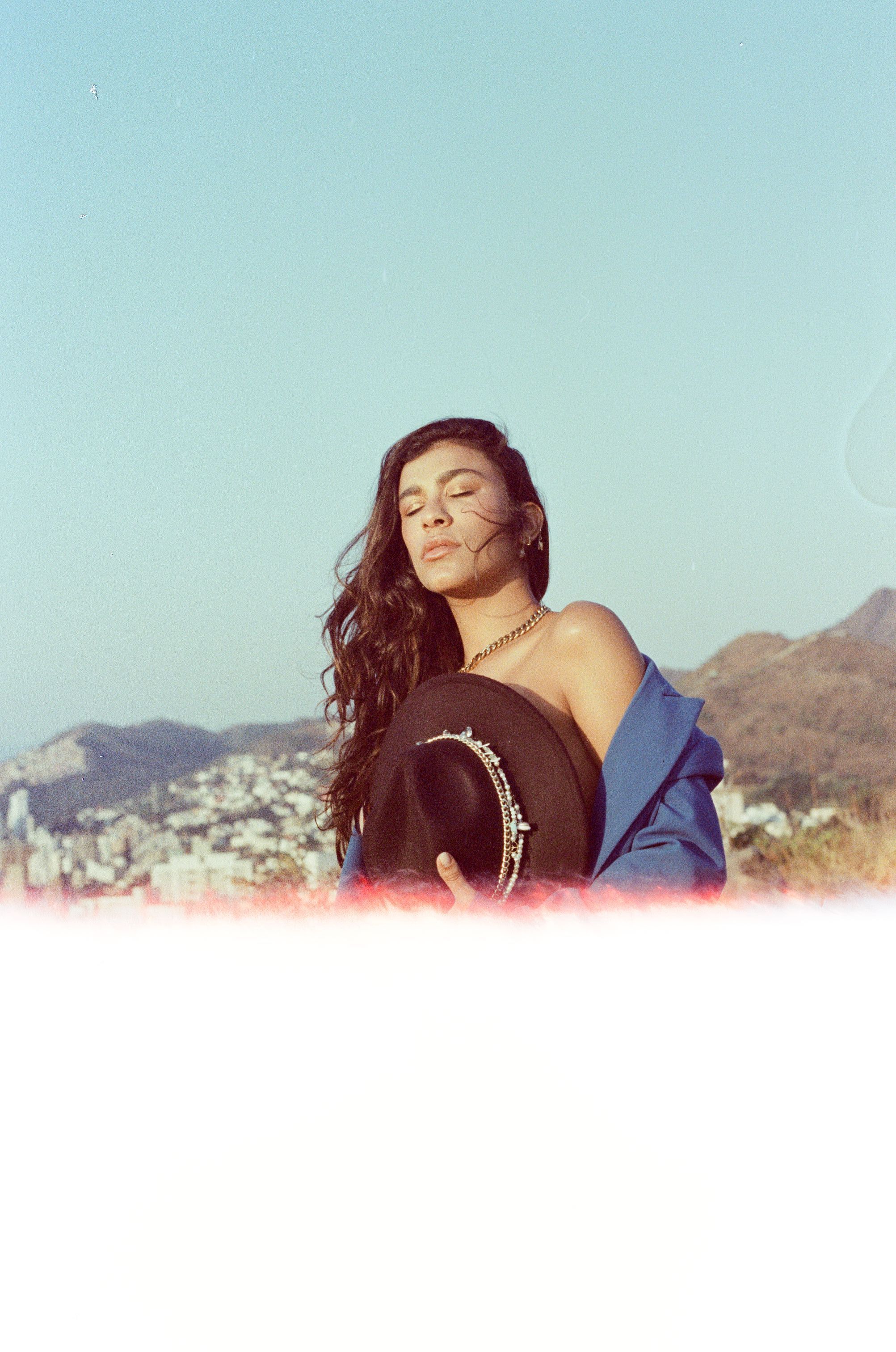 Woman with long hair and bare shoulders holding a black hat outdoors against a scenic backdrop.
