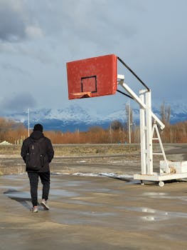 A solitary man walks towards a basketball hoop on a cloudy day with mountains in the background.