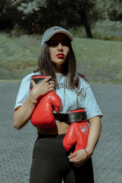 Free Young female boxer in a cap and grey shirt holding red gloves outdoors in sunlight. Stock Photo