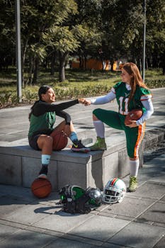 Two women in athletic gear share a fist bump, surrounded by basketball and football equipment outdoors.