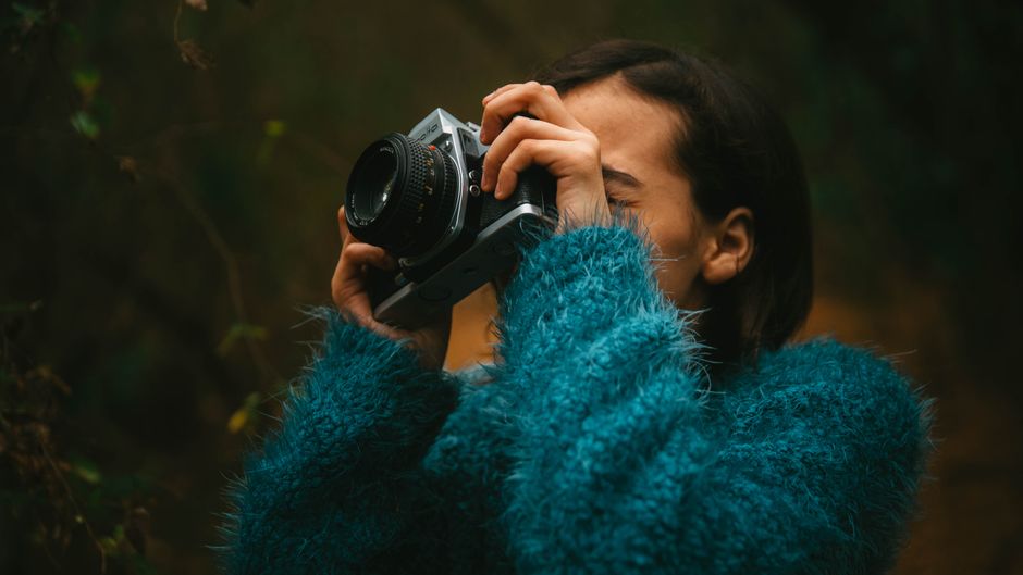 A woman in a vintage blue sweater takes photos outdoors using an analog camera.