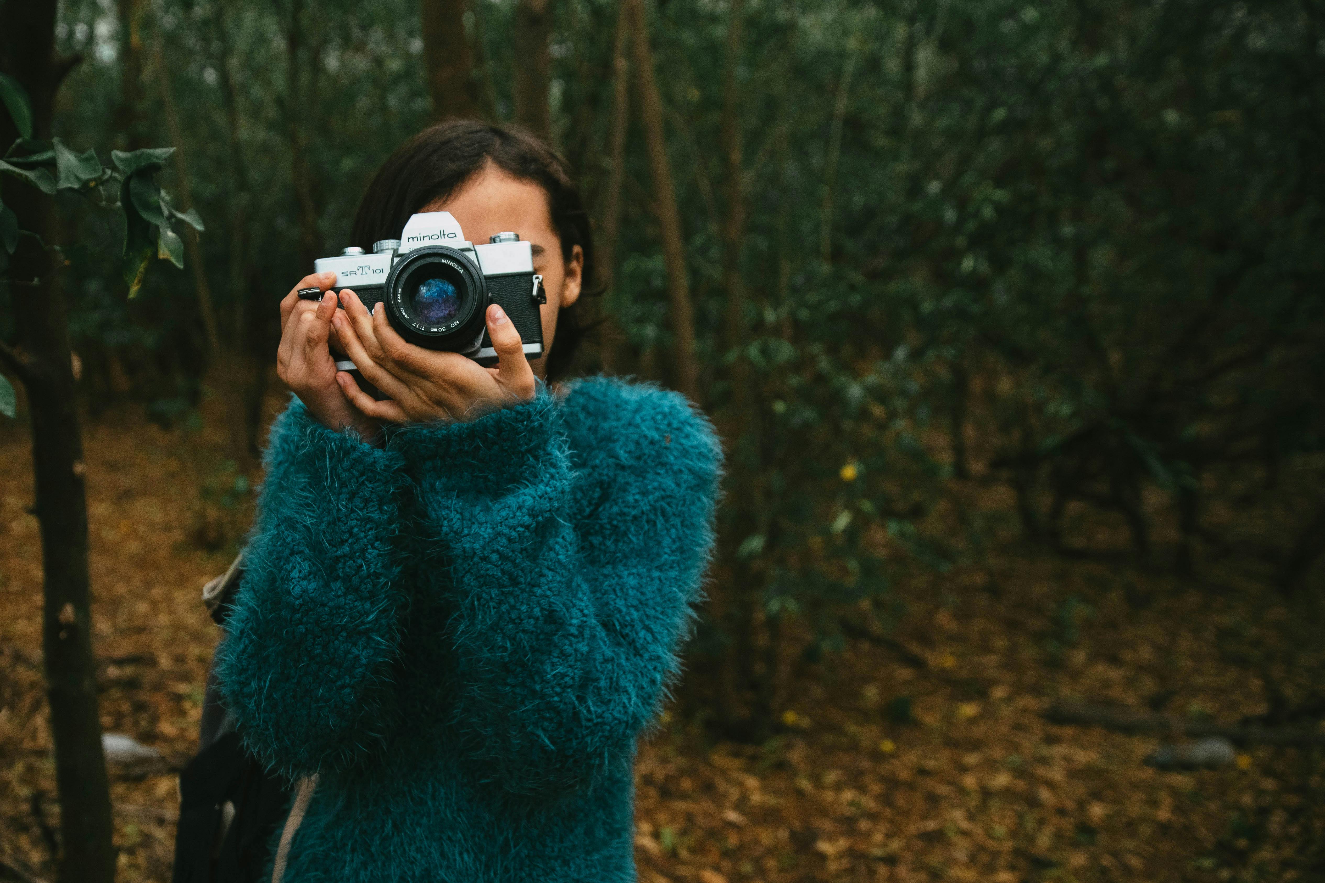 Free A woman in a teal sweater holds a vintage camera in a forest setting, capturing nature. Stock Photo