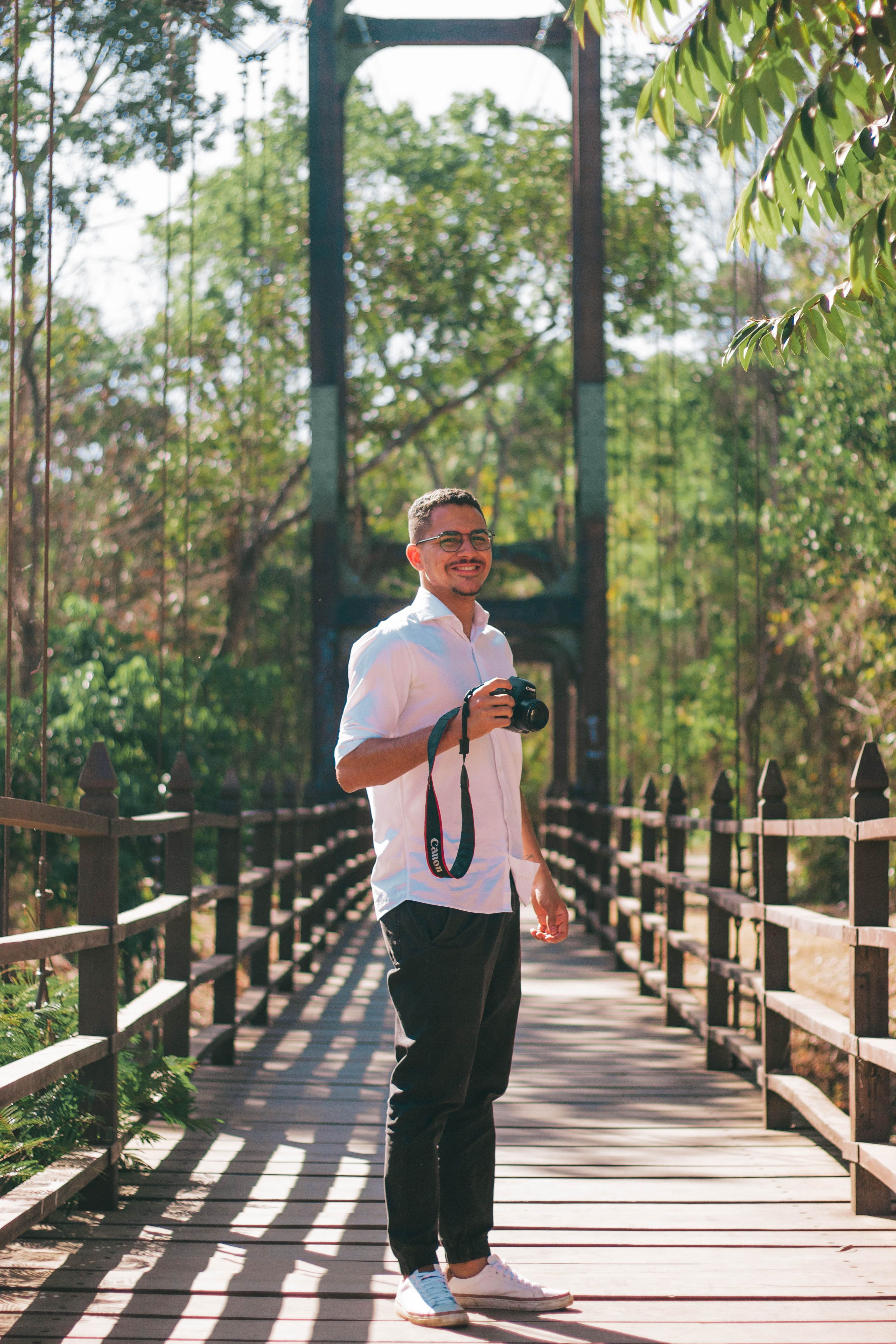 Smiling man holding camera on a wooden bridge surrounded by nature