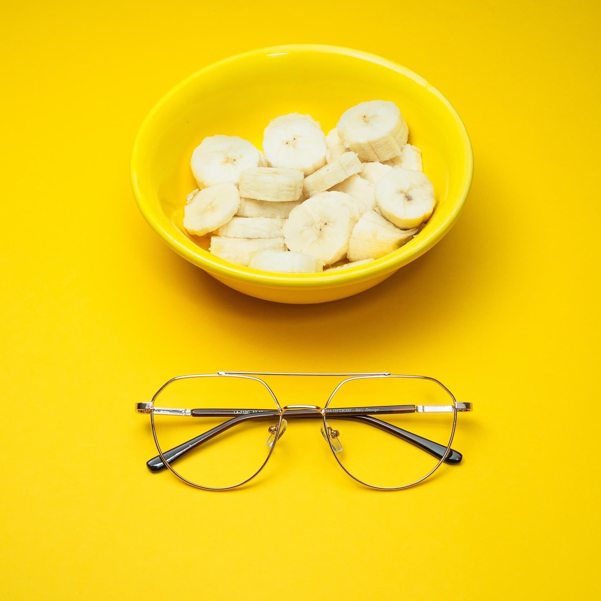 A vibrant still life photo featuring banana slices in a yellow bowl with eyeglasses on a matching yellow background.