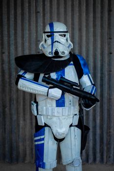 Stormtrooper cosplay costume with blue accents holding a blaster in front of a corrugated metal wall.