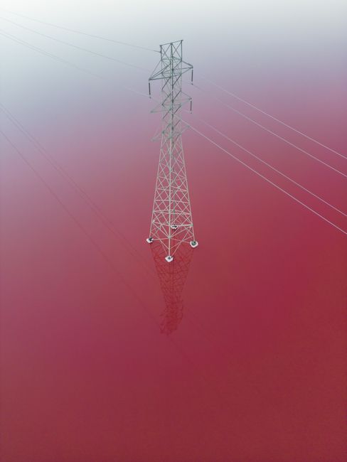 Dramatic aerial shot of an electricity tower reflected in vibrant pink water.