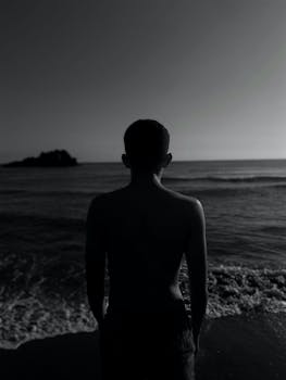 Black and white photo of a shirtless man standing at the shore, capturing the serene ocean view.