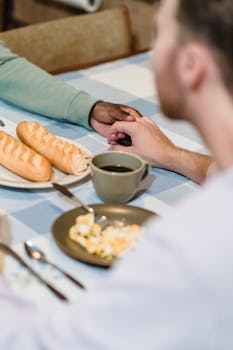 A couple shares a breakfast table holding hands, featuring bread and coffee.