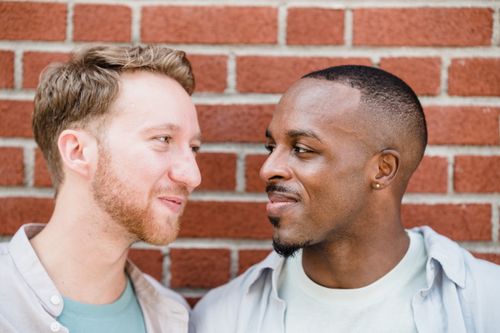 Free A happy gay couple smiling at each other, standing close against a brick wall outdoors. Stock Photo