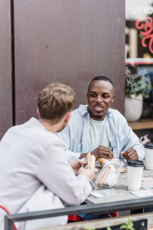 Free Two friends chatting and eating sandwiches at an outdoor cafe table. Stock Photo