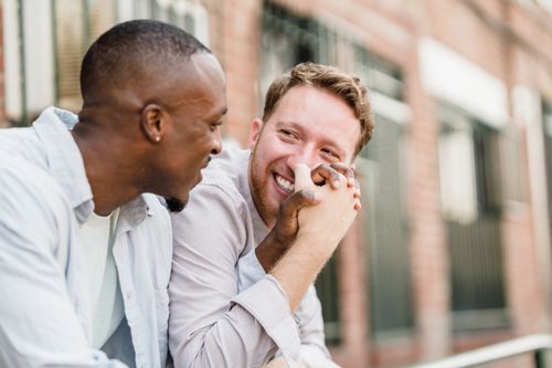 Free Happy gay couple smiling and holding hands while sitting outdoors, sharing a joyful moment together. Stock Photo