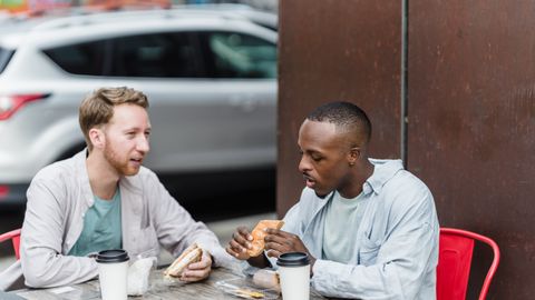 Two men having lunch outdoors at a table, enjoying sandwiches and drinks.
