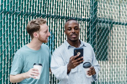 Free Two men enjoying coffee and looking at a smartphone outdoors against a fence. Stock Photo