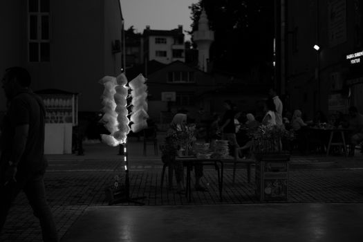 Black and white urban market scene at night with cotton candy display and people sitting.