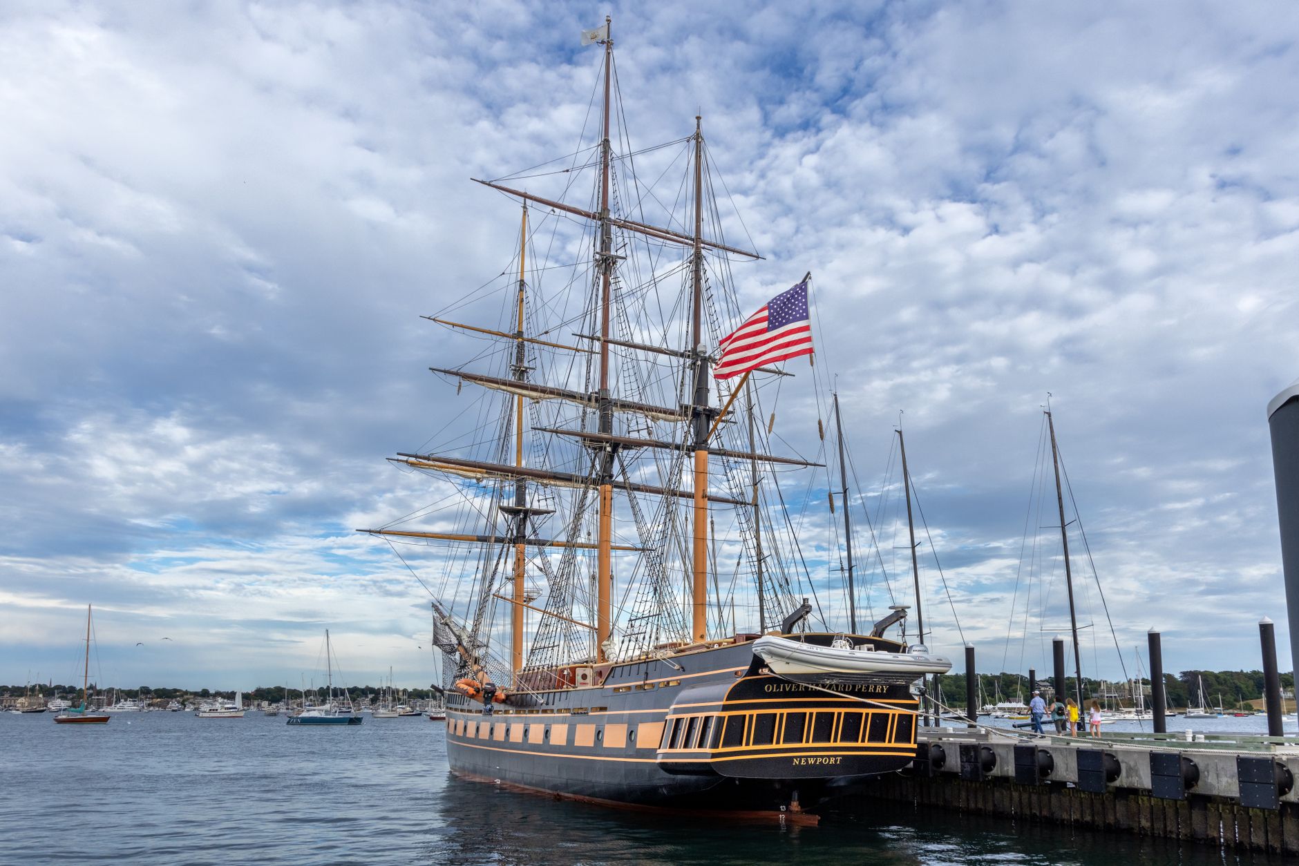 Tall ship with American flag docked at Newport Harbor under a cloudy sky.
