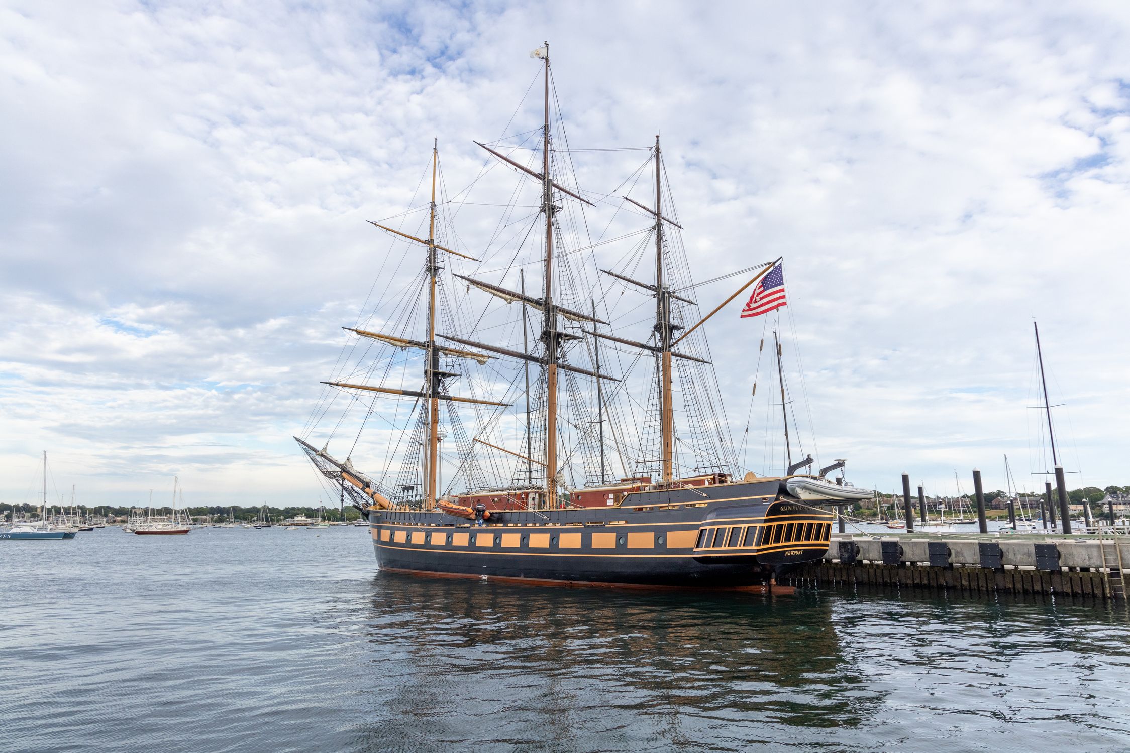 A majestic historic ship with an American flag docked in Newport, RI harbor, under a cloudy sky.