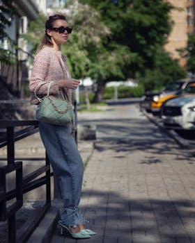 Stylish woman in sunglasses posing with a green bag and denim pants on a sunny sidewalk.