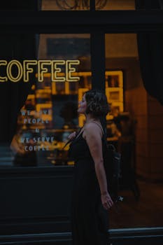 A woman in a black dress walks past a cozy coffee shop at night.