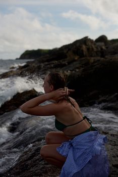 A woman in a swimsuit sits on a rocky shore adjusting her hair with waves crashing nearby.