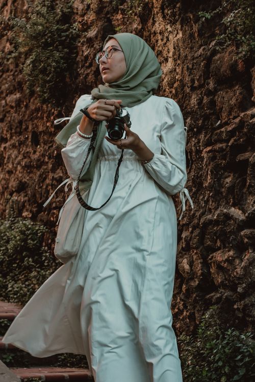 Free A woman in a white dress and hijab holding a camera against a rustic stone wall in Istanbul. Stock Photo