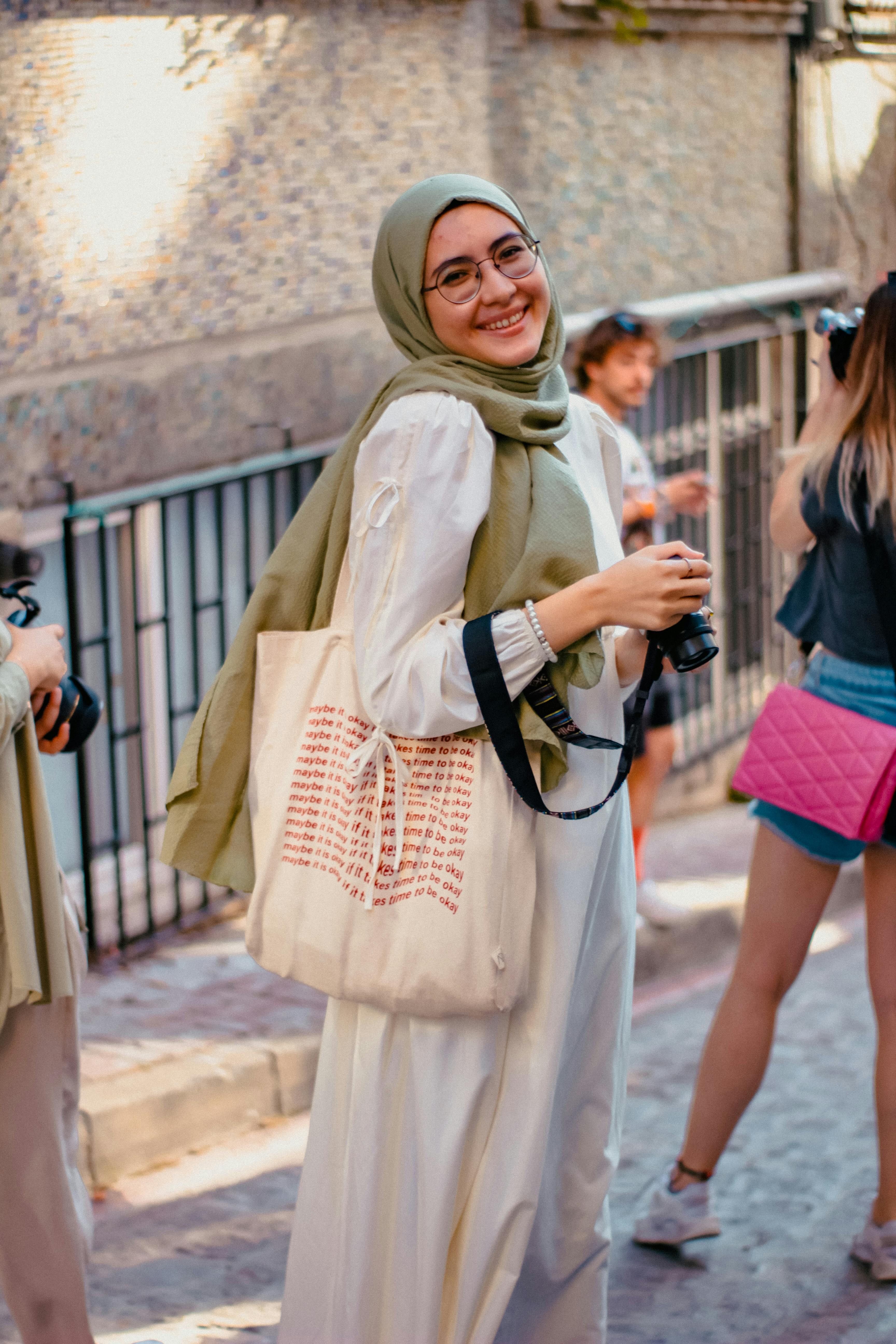 young woman smiling in white long sleeve dress and beige hijab holding a digital camera