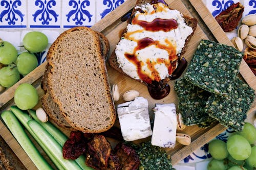 Top view of wooden cutting board with fresh cucumber slices and black bread loafs with soft tofu cheese near dry seaweed and crunchy pistachios near pineapple guava