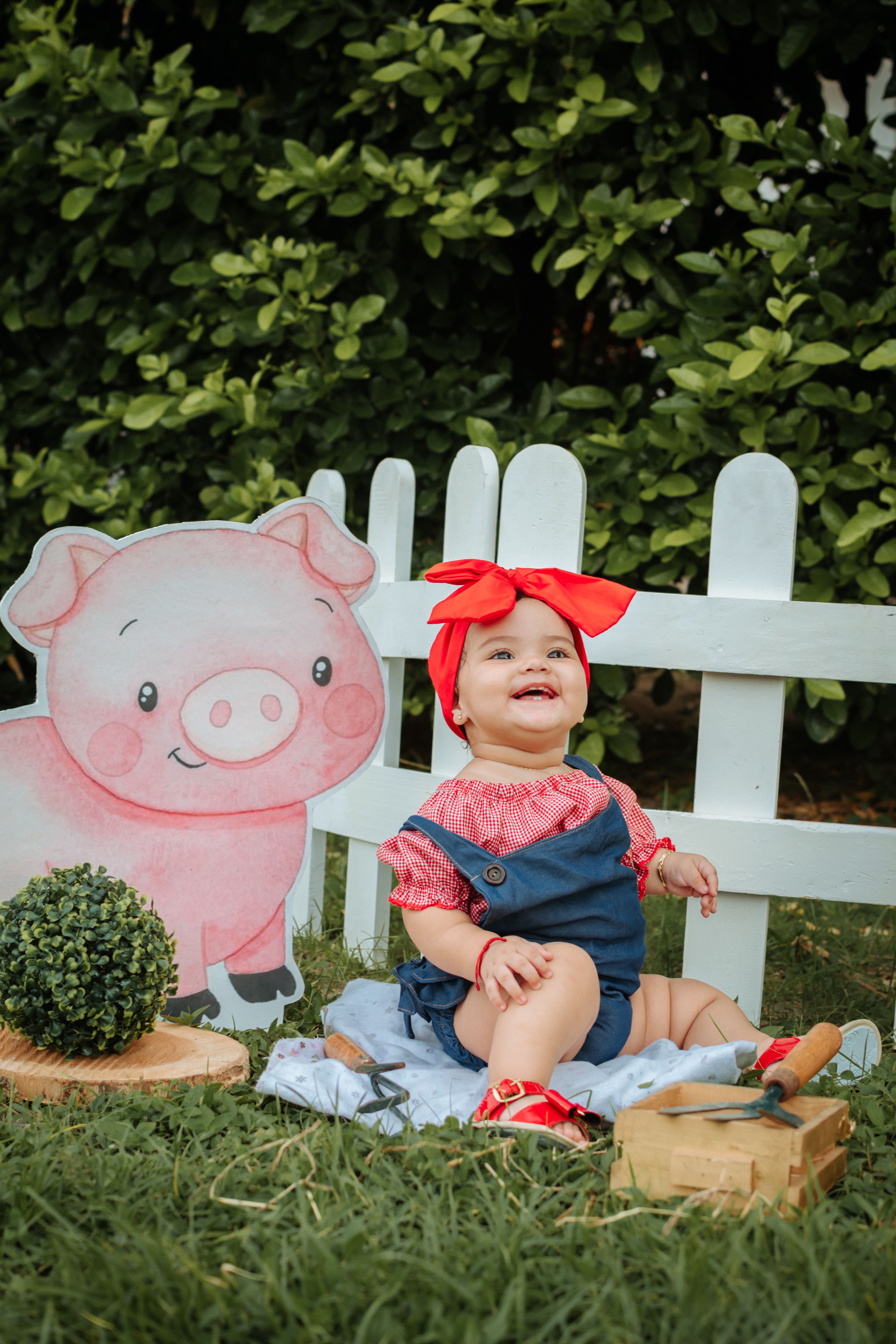 A joyful baby sitting next to a cartoon pig and a white picket fence outdoors.