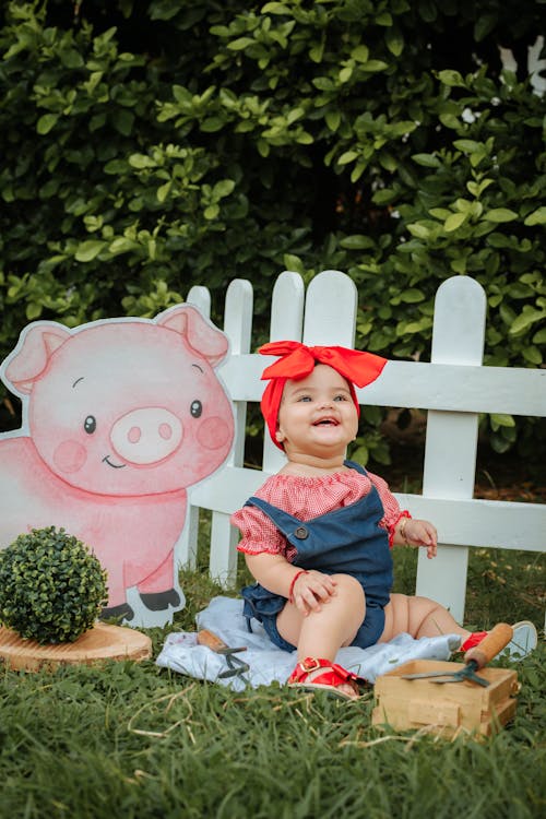 Baby Sitting on Grass by White Fence and Cutout of Cartoon Piglet