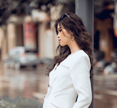 Stylish woman with curly hair poses in a white outfit against an urban backdrop.