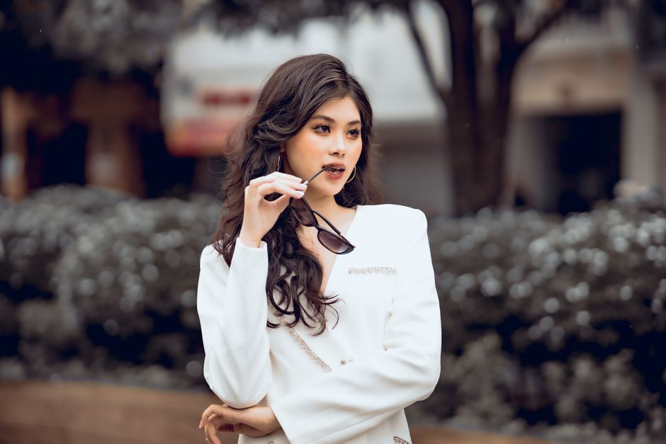 Stylish woman with curly hair poses confidently in white clothing, holding sunglasses.