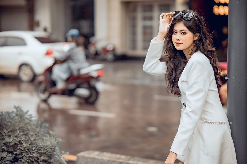 Fashionable woman in white attire posing on a rainy day street with blurred urban background.