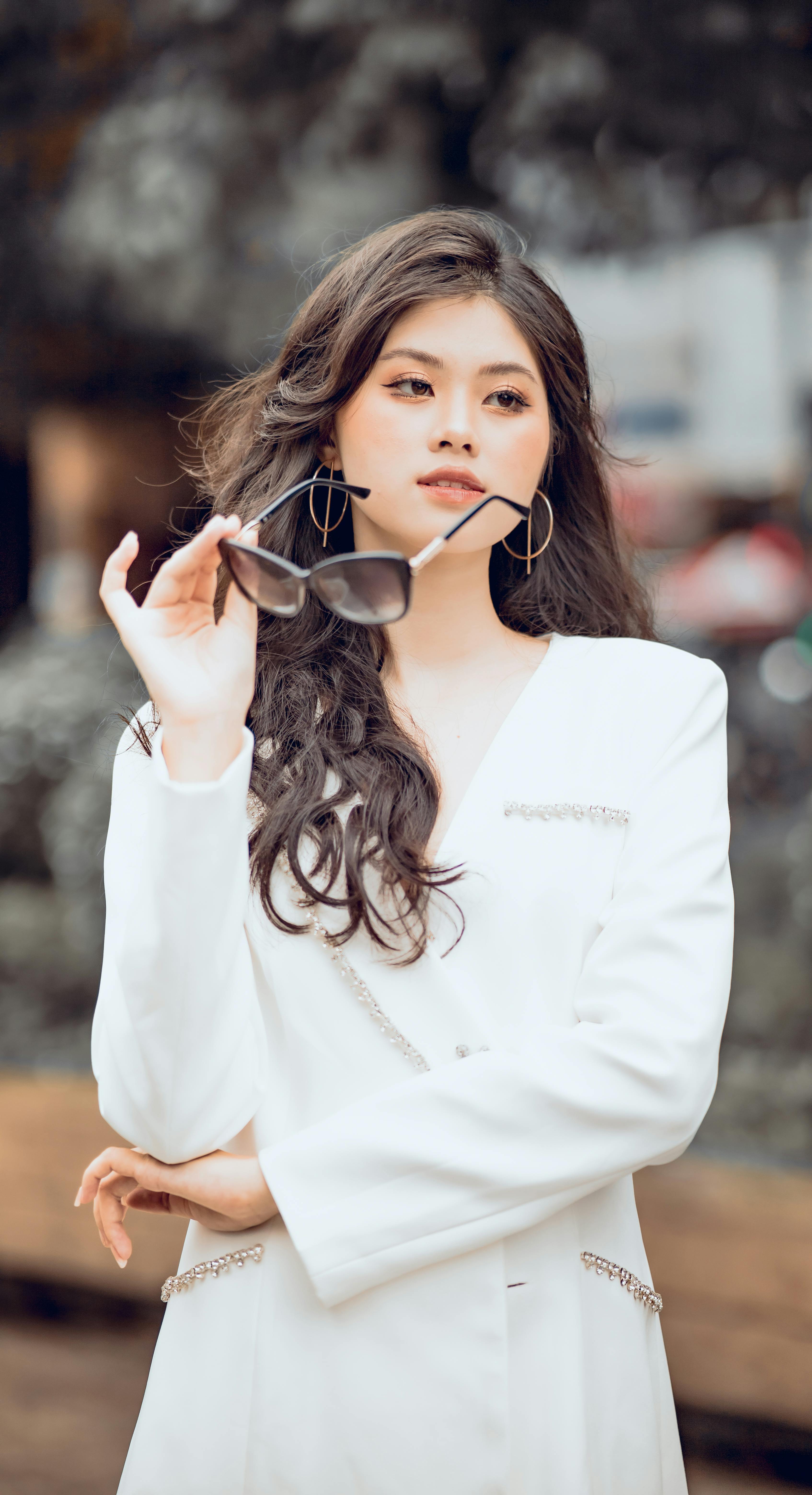 Asian woman with curly hair posing outdoors in white dress with sunglasses, embodying elegance and style.