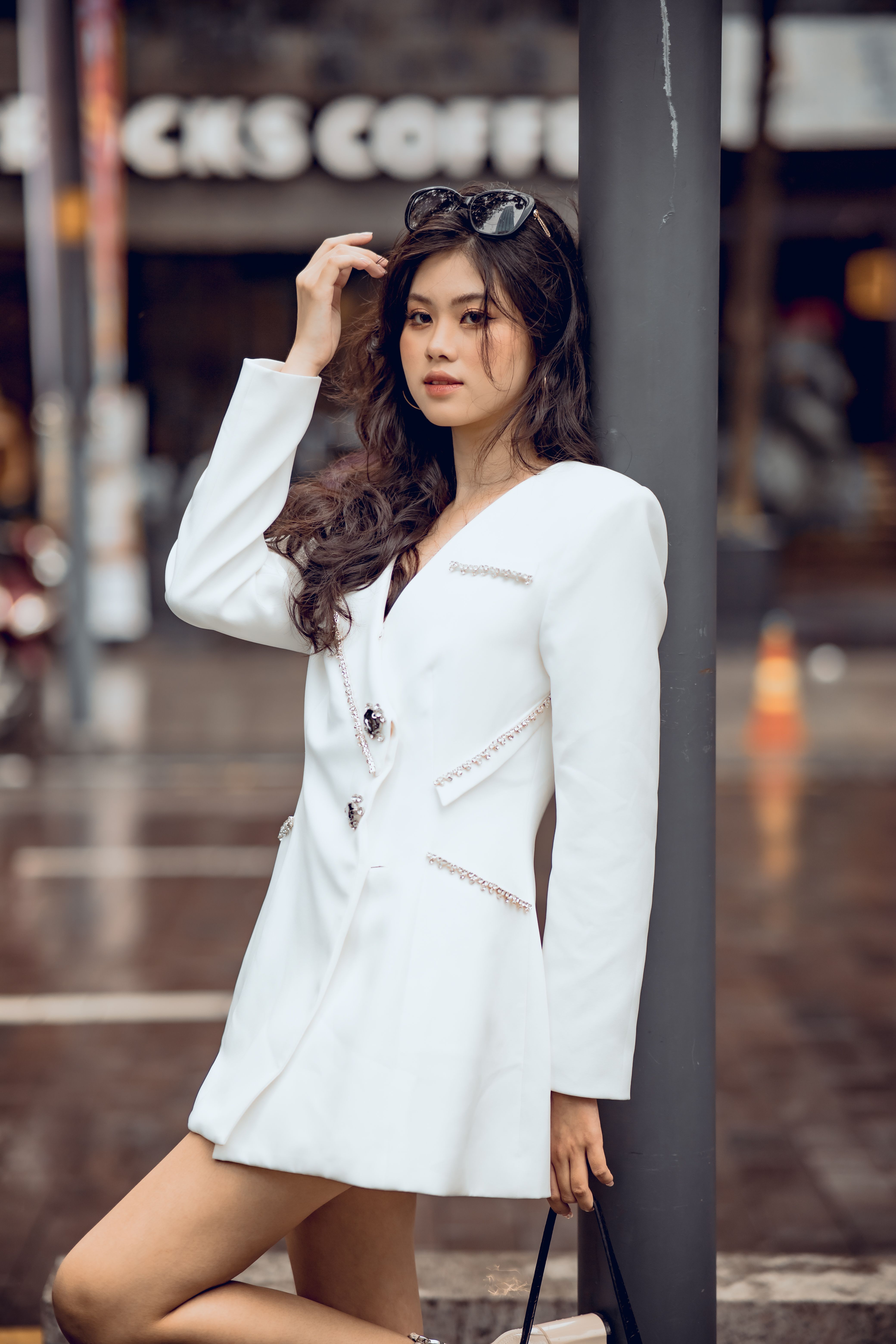Stylish Asian woman in white dress posing in urban setting with blurred background.